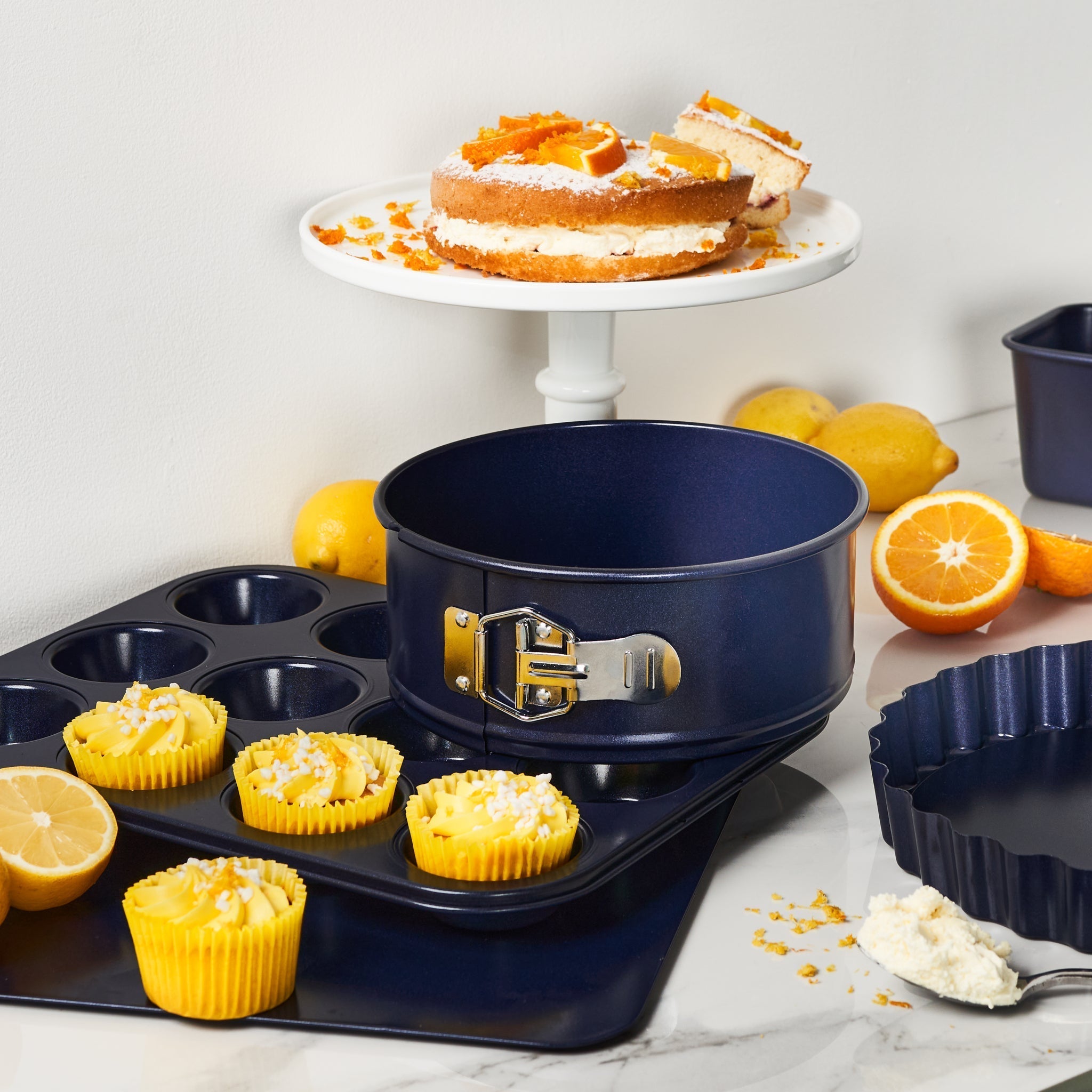 A cake on a stand, lemon cupcakes in a muffin tray, a springform pan, a tart pan, and citrus fruits are arranged on a marble countertop.
