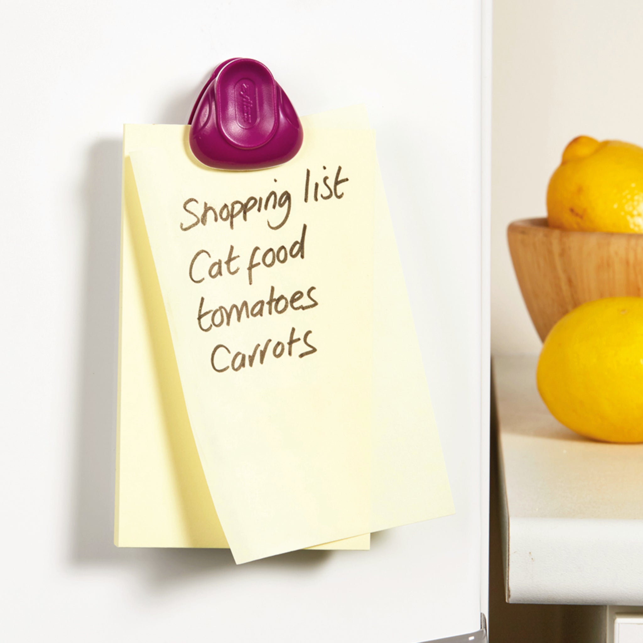 A shopping list written on a yellow sticky note is clipped to a fridge with a purple magnet. The list includes cat food, tomatoes, and carrots. Lemons in a bowl are visible on a nearby counter.