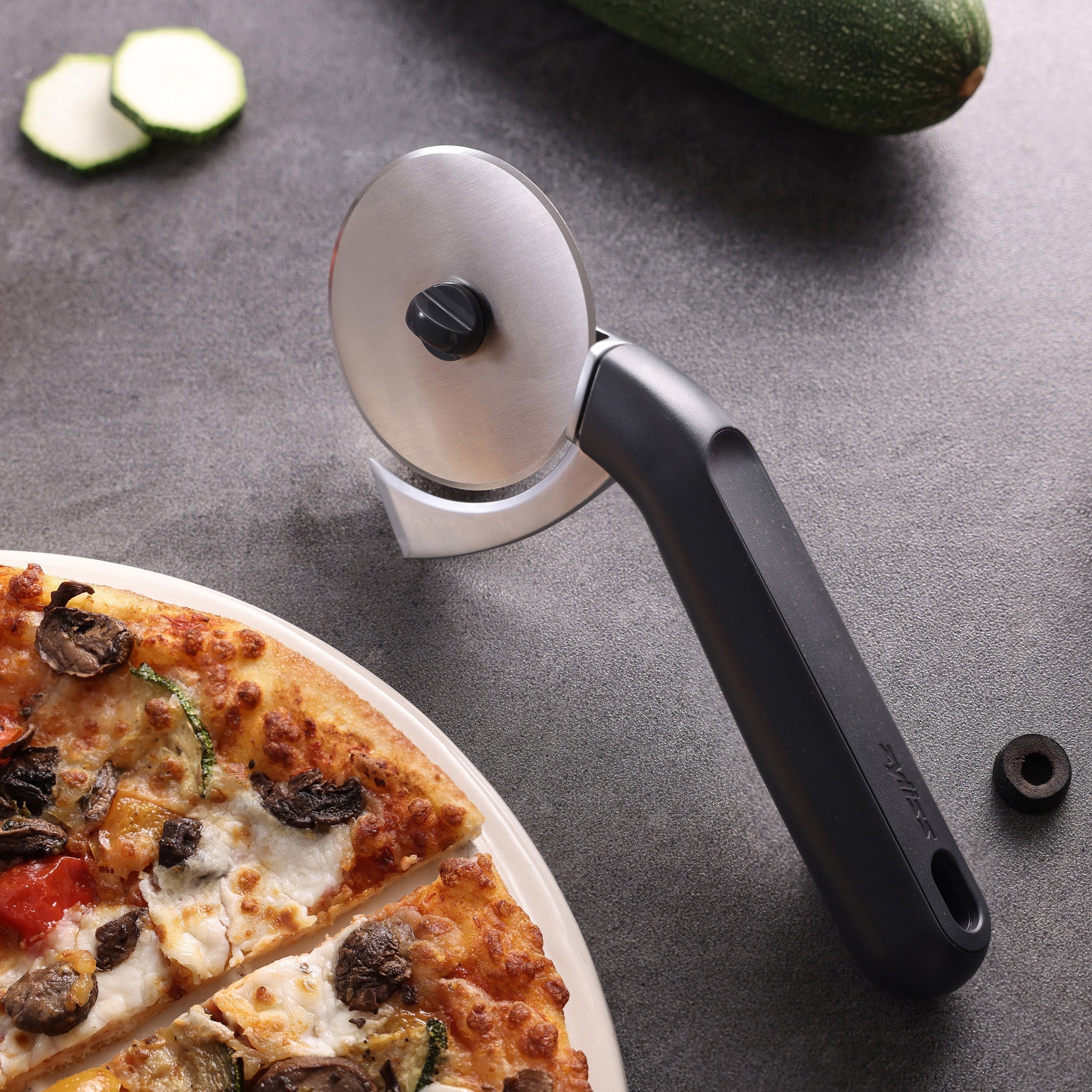A black-handled pizza cutter rests on a gray countertop next to a partially sliced pizza and cucumber slices, with a zucchini in the background.