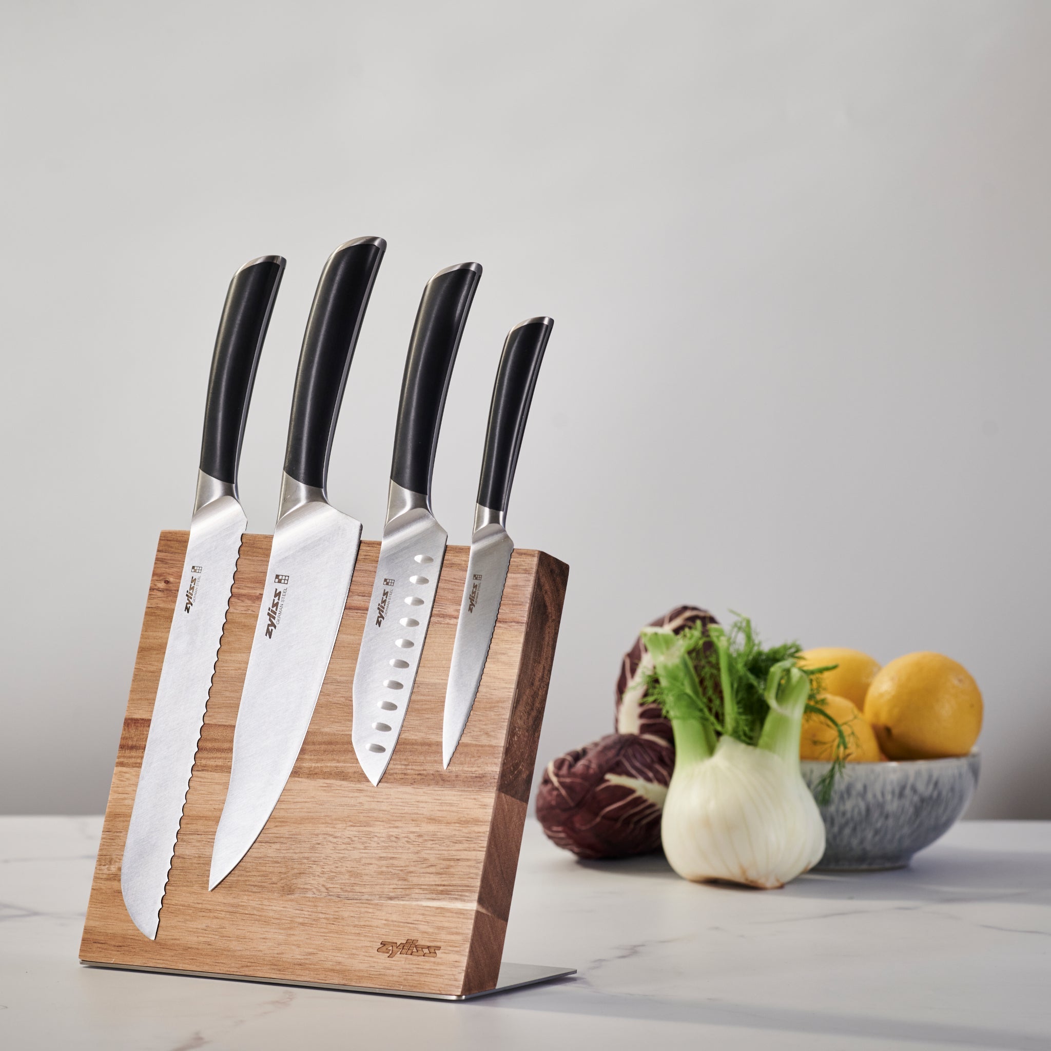 A wooden knife block holds four kitchen knives with black handles. In the background, a bowl contains lemons, fennel, and radicchio, sitting on a light countertop.