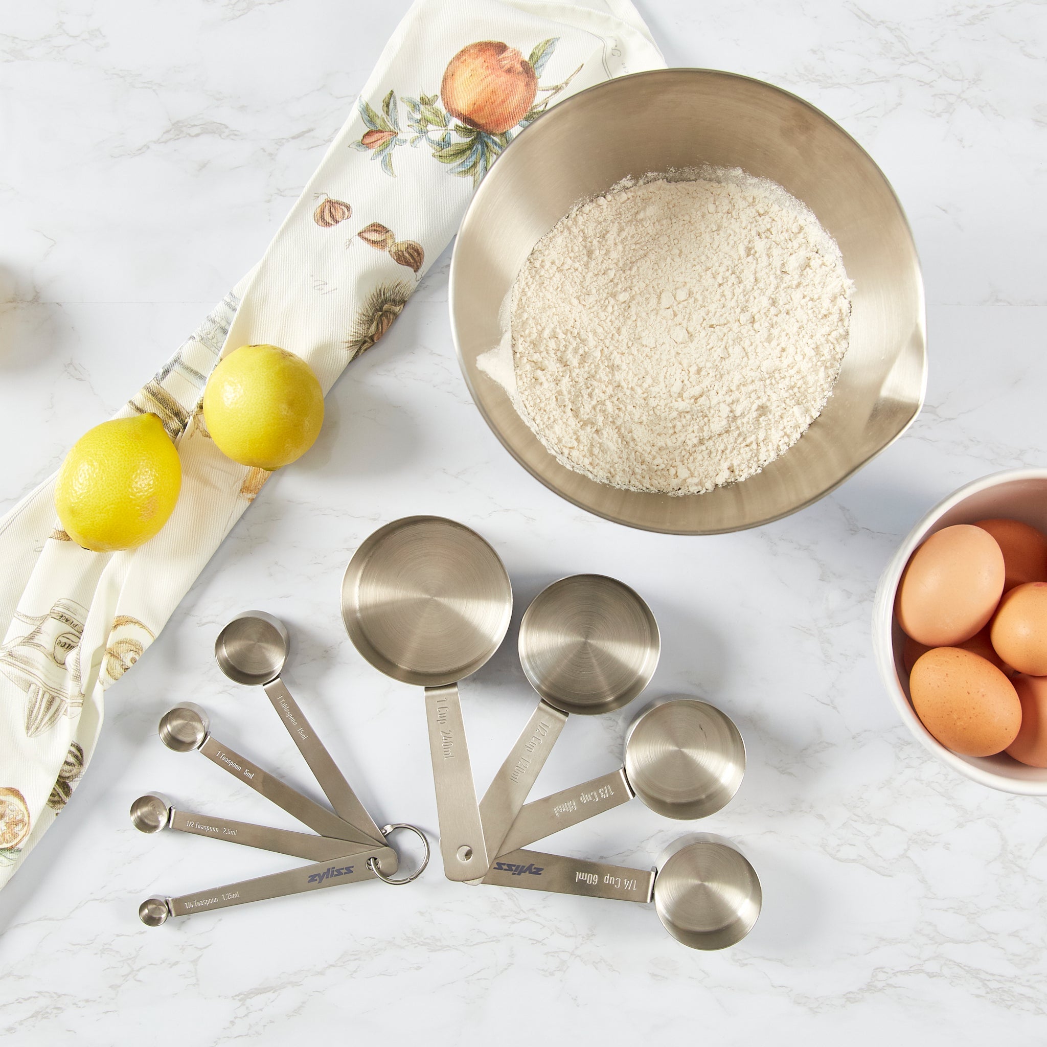 A set of metal measuring cups and spoons arranged on a marble counter with a bowl of flour, a bowl of eggs, two lemons, and a decorative kitchen towel with fruit illustrations.