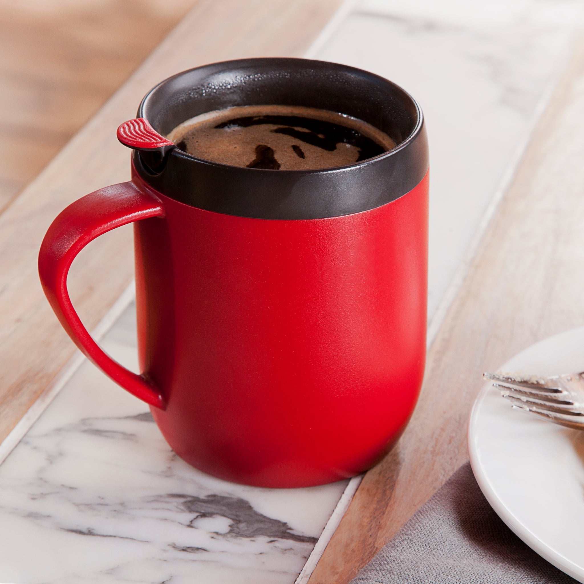 A red mug filled with coffee sits on a marble and wood table. A small red-handled spoon rests inside the mug. A plate with a fork and knife is partially visible nearby.