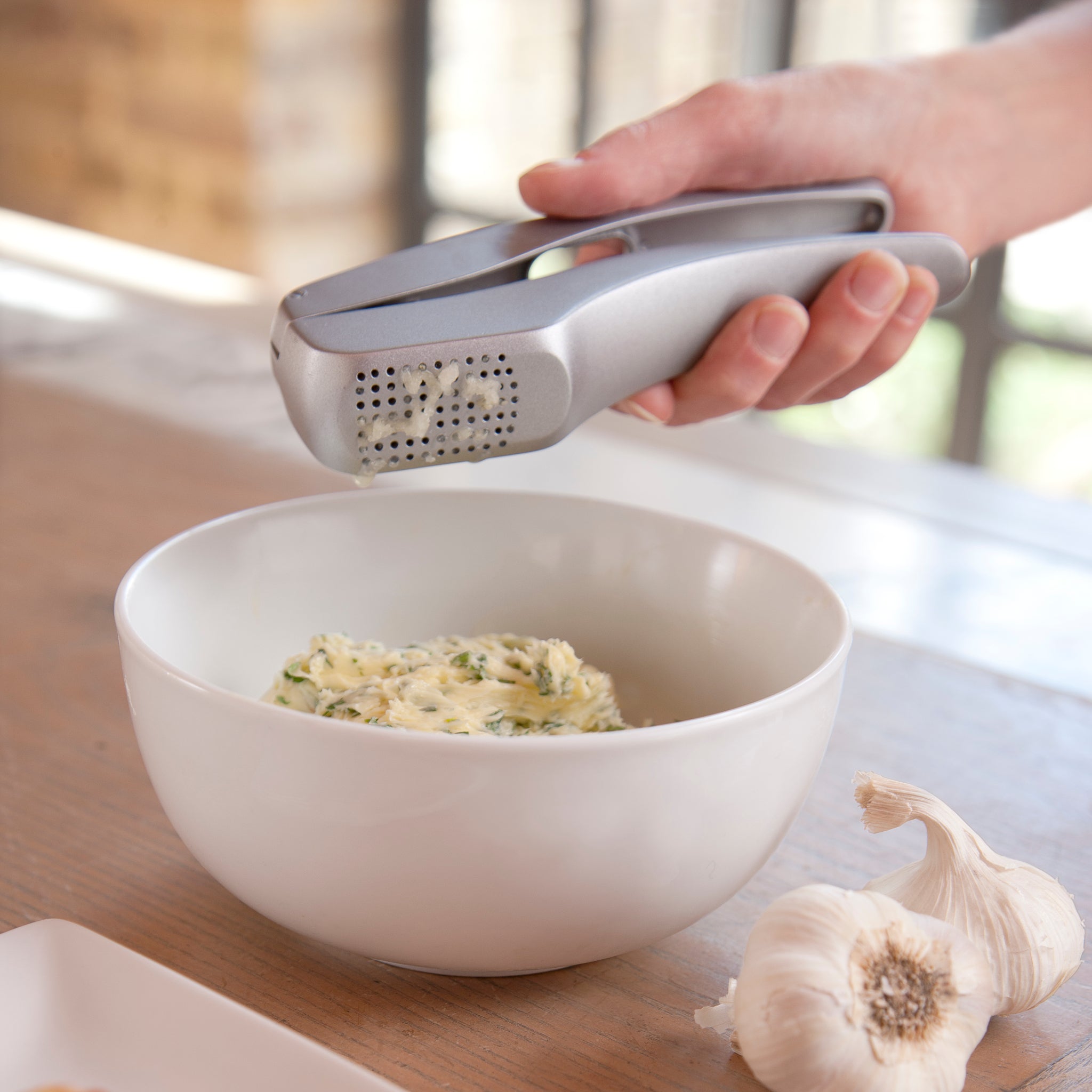 A hand squeezes a garlic press over a white bowl filled with an herbed mixture. Two garlic bulbs sit on the wooden table nearby.