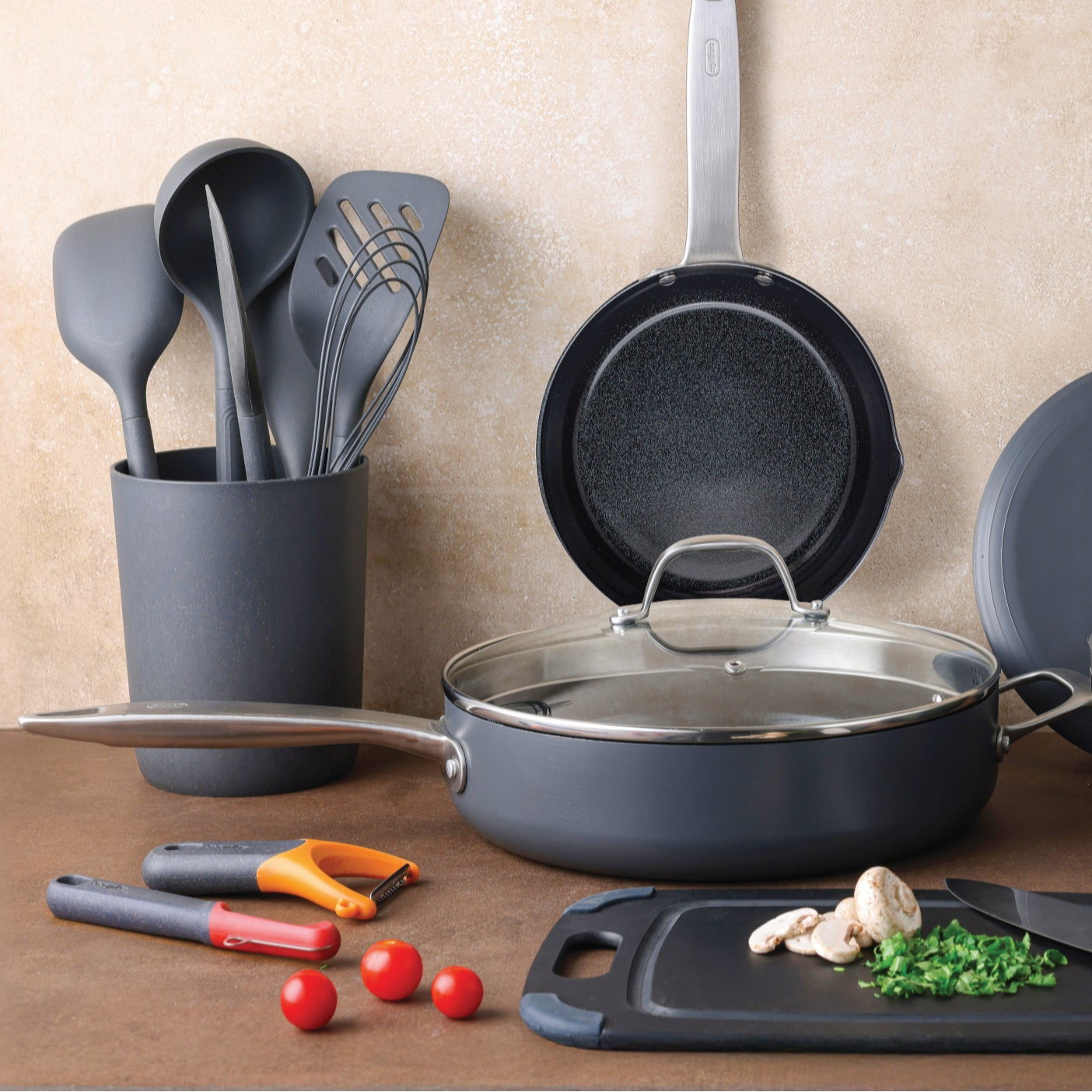 A set of gray kitchen utensils in a holder, a frying pan hanging on the wall, a lidded pan, cutting board with mushrooms, chopped herbs, tomatoes, and peelers on a wooden surface.