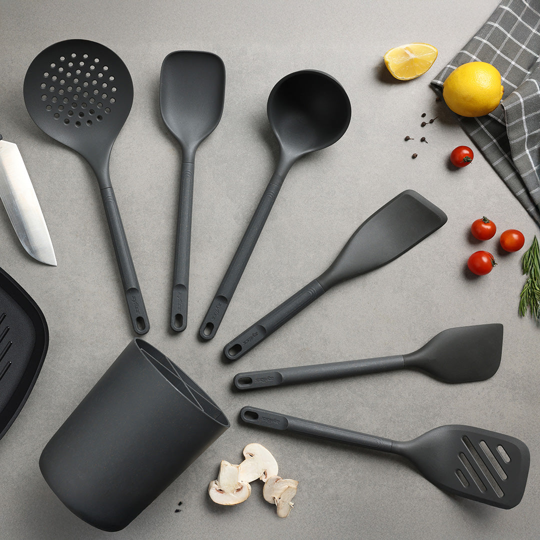 A set of six black kitchen utensils arranged on a countertop with a utensil holder, knife, mushrooms, cherry tomatoes, lemon halves, and a checkered towel nearby.
