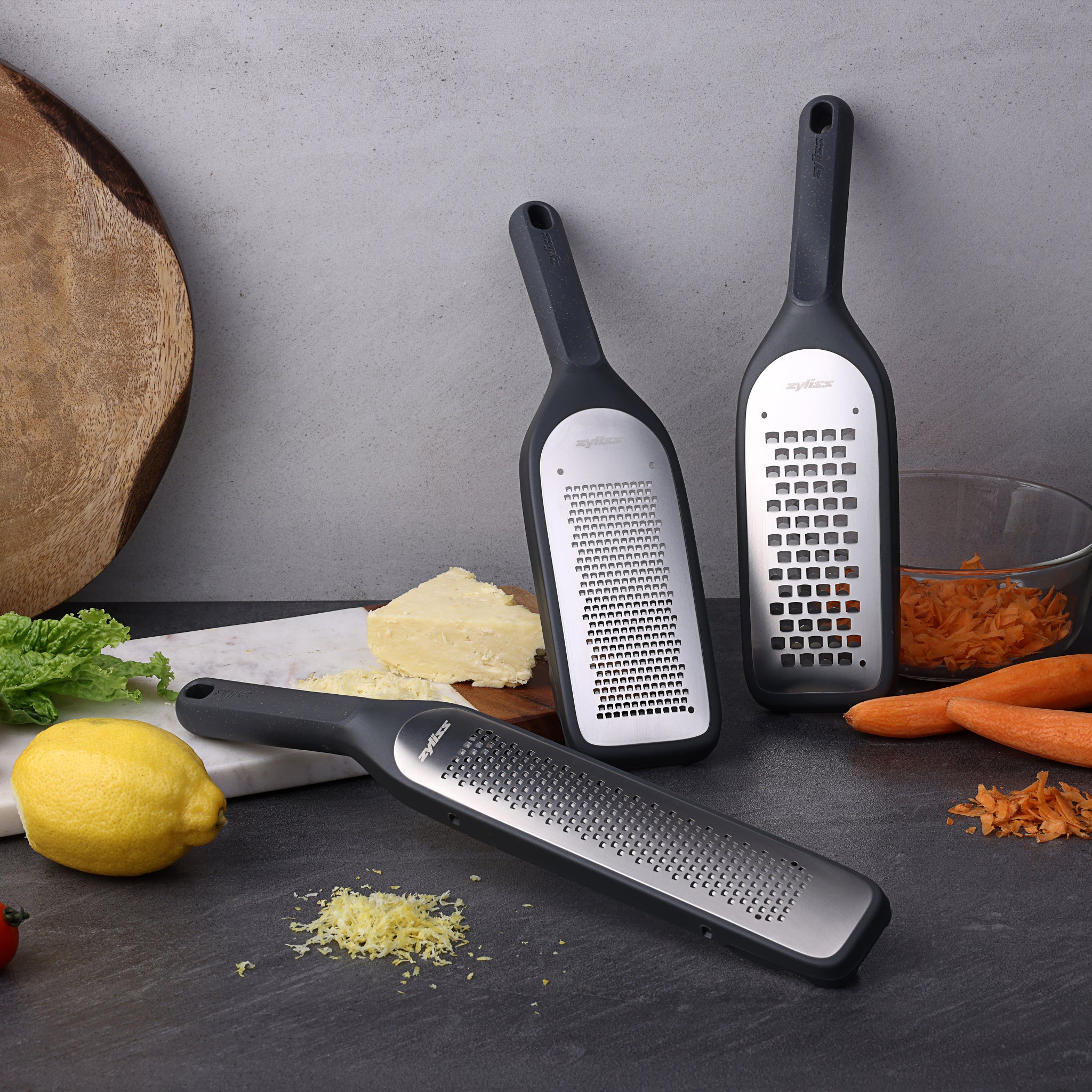 Three black-handled graters stand upright on a kitchen counter, surrounded by grated cheese, shredded carrots, whole carrots, lettuce, a lemon, and a large wooden board.