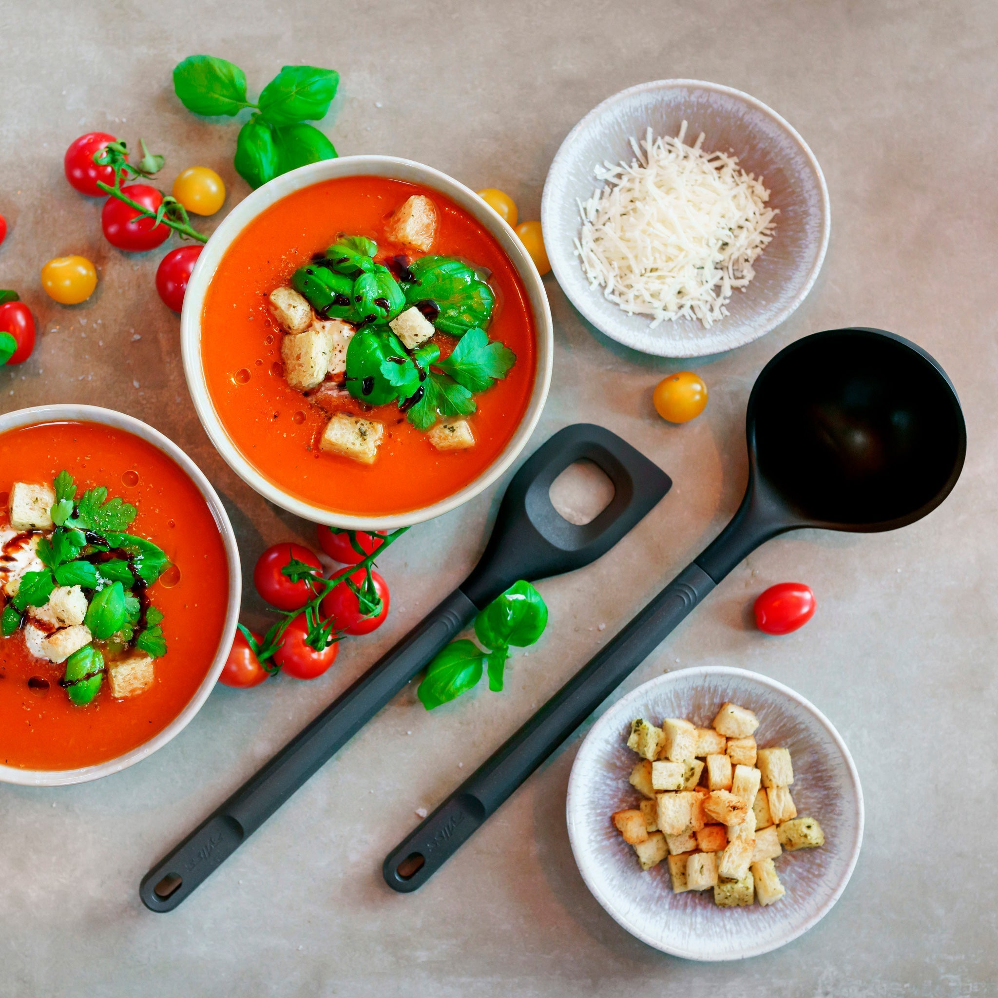 Two bowls of tomato soup garnished with parsley and croutons on a table. Surrounded by cherry tomatoes, basil, bowls of grated cheese and croutons. Two black ladles are placed between the bowls.