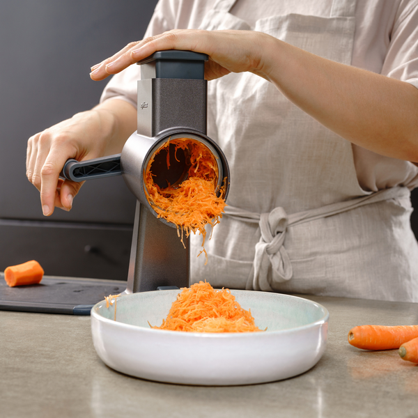 A person is shredding carrots into a bowl on a kitchen counter using the Zyliss Fine Grating Drum, with additional carrots and a cutting board nearby.