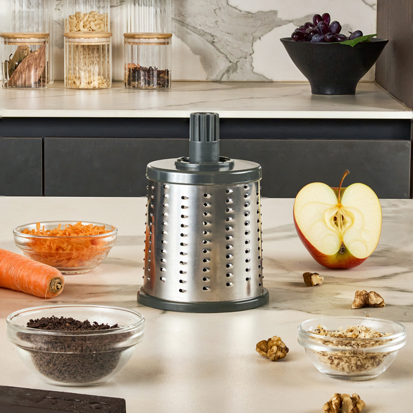 A Zyliss Fine Grating Drum stands ready for use on a kitchen counter, surrounded by shredded carrots, walnuts, apple halves, spices, and a whole carrot. In the background are jars and a fruit bowl.