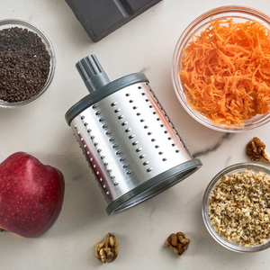 A Zyliss Fine Grating Drum takes center stage, ready for action, encircled by a red apple, a bowl of shredded carrots, a bowl of chopped walnuts, pieces of chocolate, and scattered crushed chocolate and walnuts on a light surface.