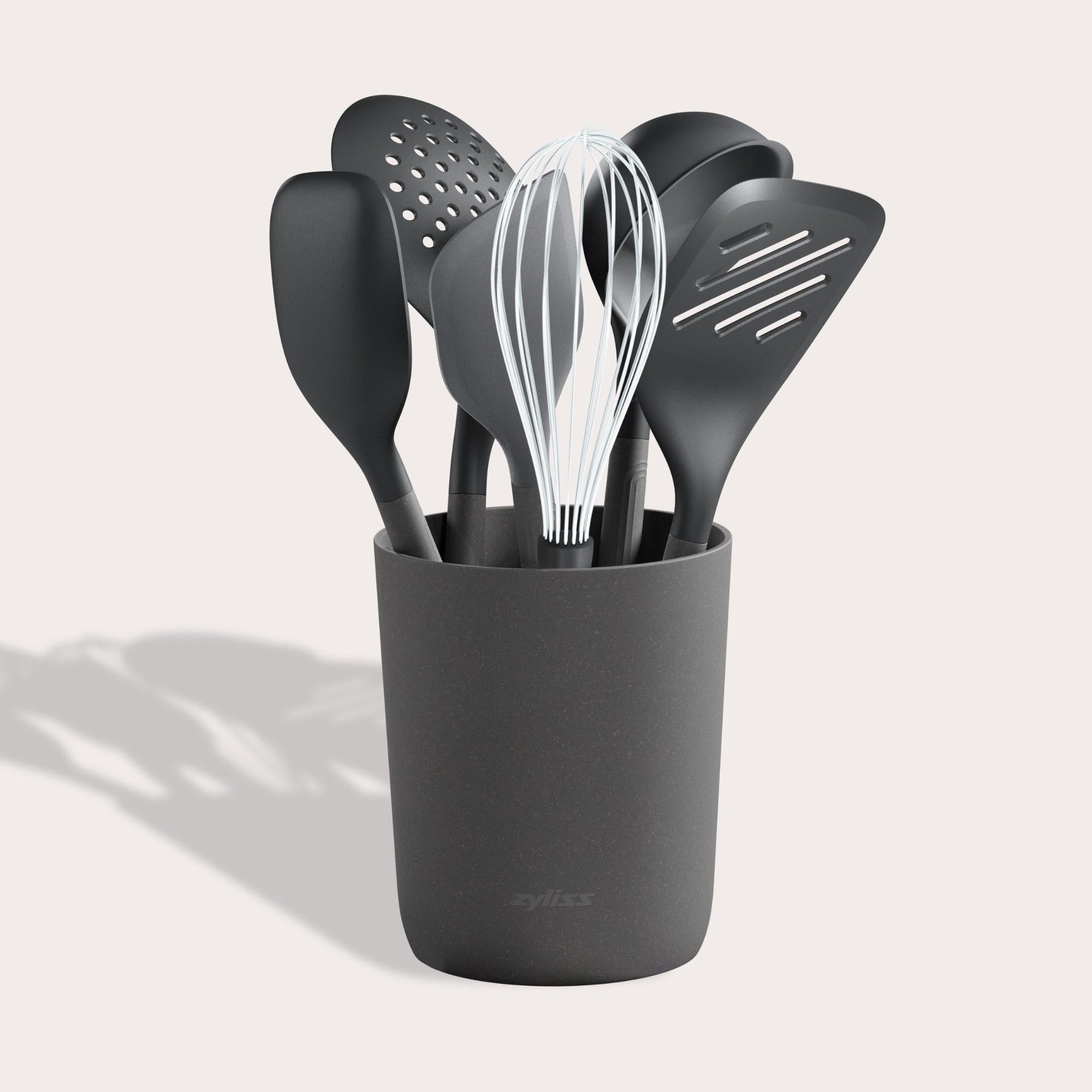 The Zyliss 7-Piece Utensil Set includes black kitchen tools—whisk, spatulas, spoon, and slotted spoon—organized in a matching container. Each features ergonomic handles and heat-resistant PBT for safe, comfortable use. 