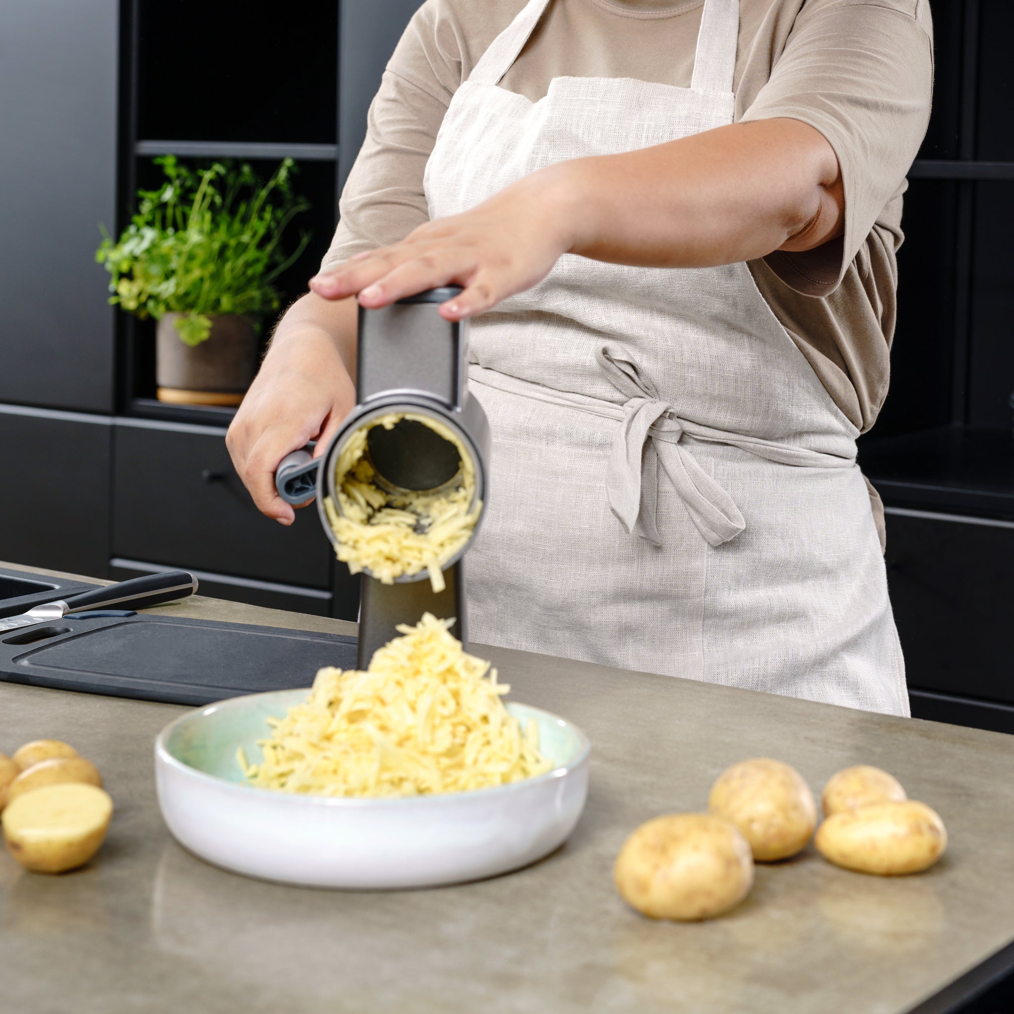 In a modern kitchen, a person uses the Zyliss Puree Drum Gourmet with its stainless steel drum to grate potatoes over a bowl. Whole and halved potatoes rest on the counter, with a potted herb adding greenery in the background. 