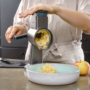 #drumgraterset Wearing an apron, someone uses the Zyliss Gourmet Drum Grater to shred apples into a light blue bowl on the kitchen counter, with a whole apple nearby.