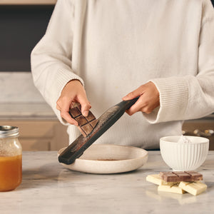 Wearing a white sweater, someone uses the Zyliss Rasp Grater with an ultra-sharp blade to grate chocolate over a shallow bowl on a marble countertop, with chocolate pieces, a jar, and white bowl nearby. 