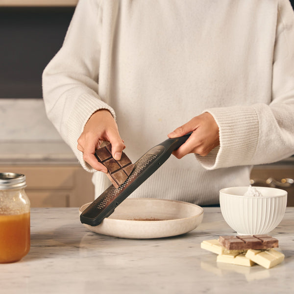 Wearing a white sweater, someone uses the Zyliss Rasp Grater with an ultra-sharp blade to grate chocolate over a shallow bowl on a marble countertop, with chocolate pieces, a jar, and white bowl nearby. 