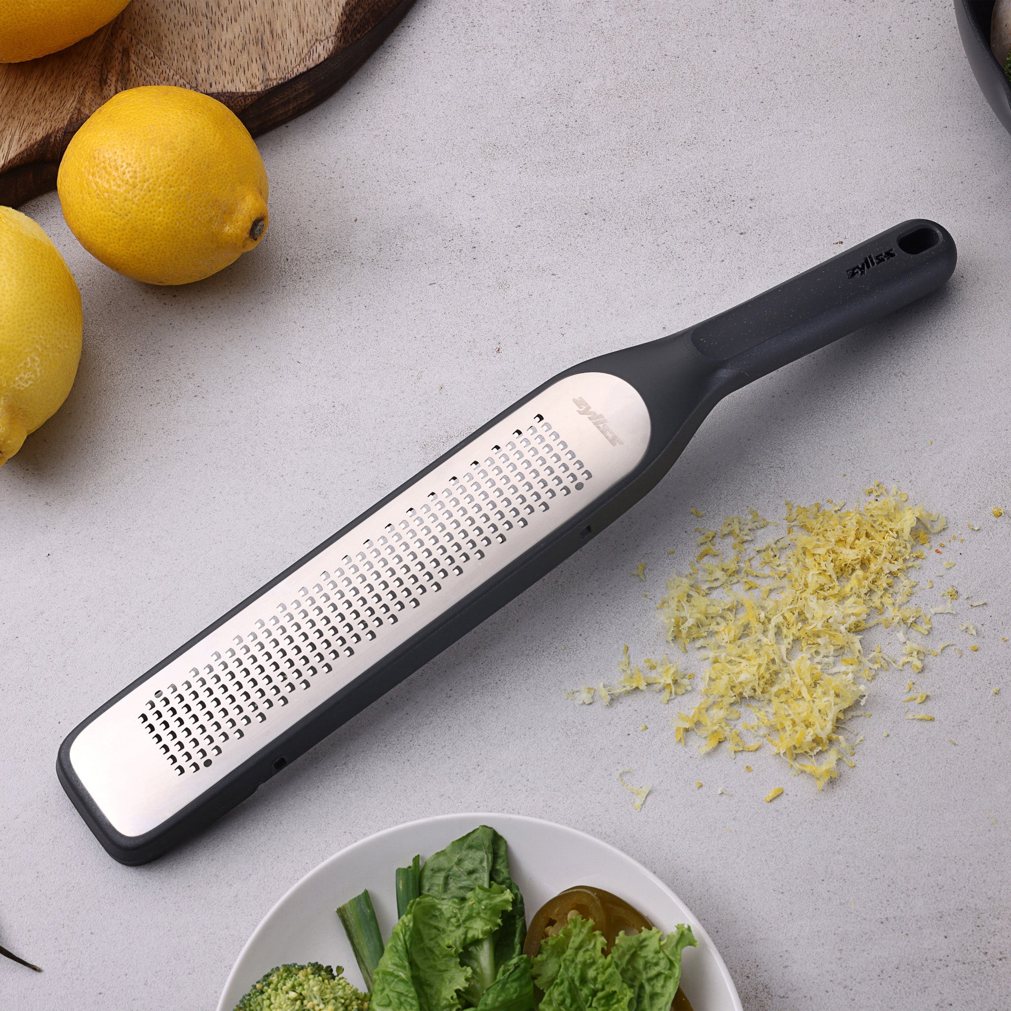 A Zyliss Rasp Grater with a black handle sits on a light countertop beside fresh lemon zest, whole lemons, and a plate of green vegetables. 