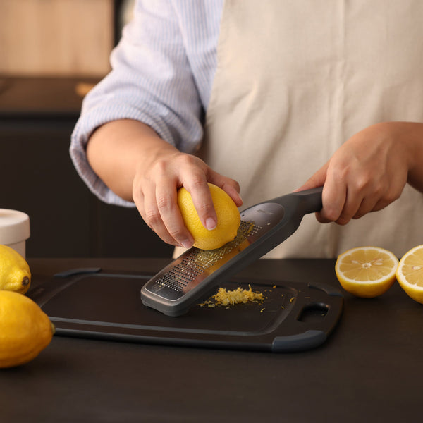 A person wearing a beige apron uses the ultra-sharp Zyliss Fine Grater to zest a lemon over a black cutting board, surrounded by lemons on a kitchen counter—a true sustainable kitchen essential. 