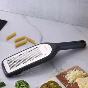 The Zyliss Fine Grater, featuring an ultra-sharp blade and black handle, sits on a kitchen counter with uncooked pasta, broccoli, jalapeño slices, and shredded cheese—a sustainable essential for easy meal prep. 