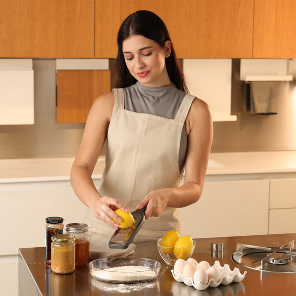 A woman in an apron smiles as she uses the Zyliss Fine Grater to zest a lemon over a bowl of flour at the kitchen counter, surrounded by eggs, jars, lemons, and baking tools. 