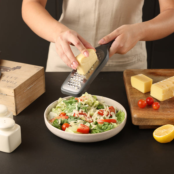 Using the Zyliss Coarse Graters ultra-sharp blade, a person grates cheese over a salad with lettuce and cherry tomatoes. Cheese blocks, a halved lemon, and tomatoes sit on a wooden board beside salt and pepper shakers. 