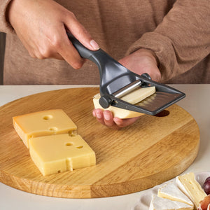 A person uses the Zyliss Dial & Slice Cheese Slicer, featuring an adjustable stainless steel blade, to cut cheese slices from a block on a round wooden cutting board while other cheese pieces are nearby.