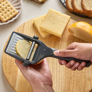 Using the Zyliss Dial & Slice Cheese Slicer with a stainless steel blade, a person cuts cheese on a wooden board. Nearby, sliced cheese, crackers, and bread are visible.