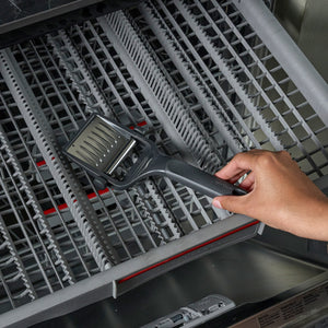 A hand places the Zyliss Dial & Slice Cheese Slicer, featuring a stainless steel grater head, on the top rack of a dishwasher among gray prongs, showing that it’s dishwasher safe.