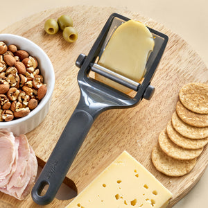 The Zyliss Dial & Slice Cheese Slicer sits on a wooden board with a cheese slice, alongside a cheese block, round crackers, sliced deli meat, mixed nuts in a bowl, and green olives.