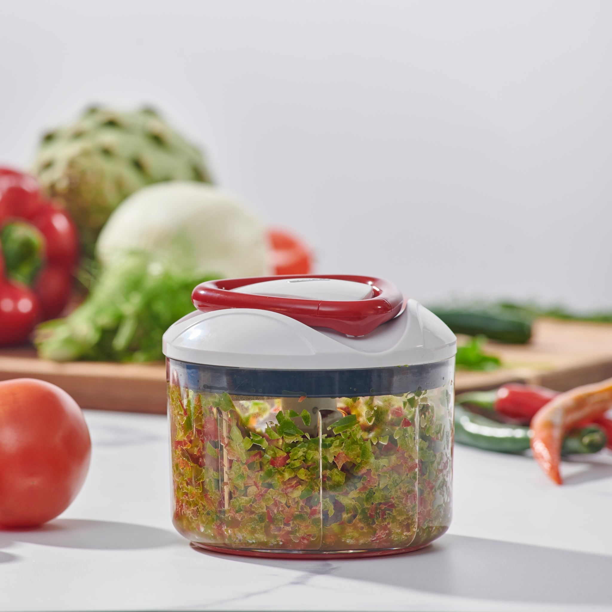 The Zyliss Easy Pull Food Processor, filled with chopped vegetables, sits on a white kitchen counter. In the background, whole tomatoes, onion, and bell pepper are displayed on a wooden cutting board. 