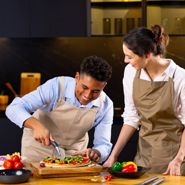 In a modern kitchen, two people in aprons smile while one slices a fresh pizza using the Zyliss Pizza & Pastry Cutter with an ergonomic handle. Colorful vegetables sit on the table, and they look like theyre enjoying cooking together. 
