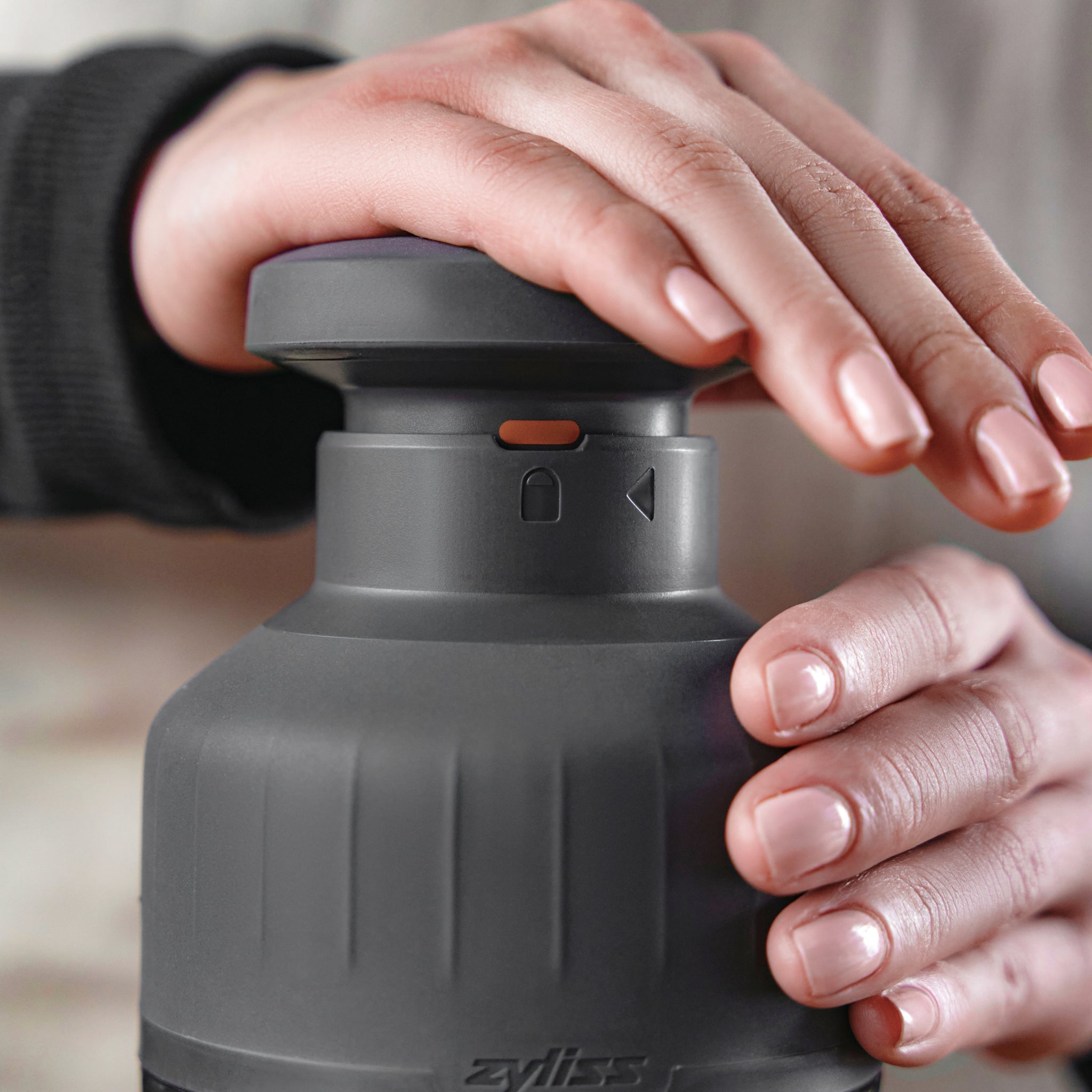 A person’s hands are shown using a gray Zyliss kitchen tool, pressing down on the top, with neatly manicured nails and a soft focus background.