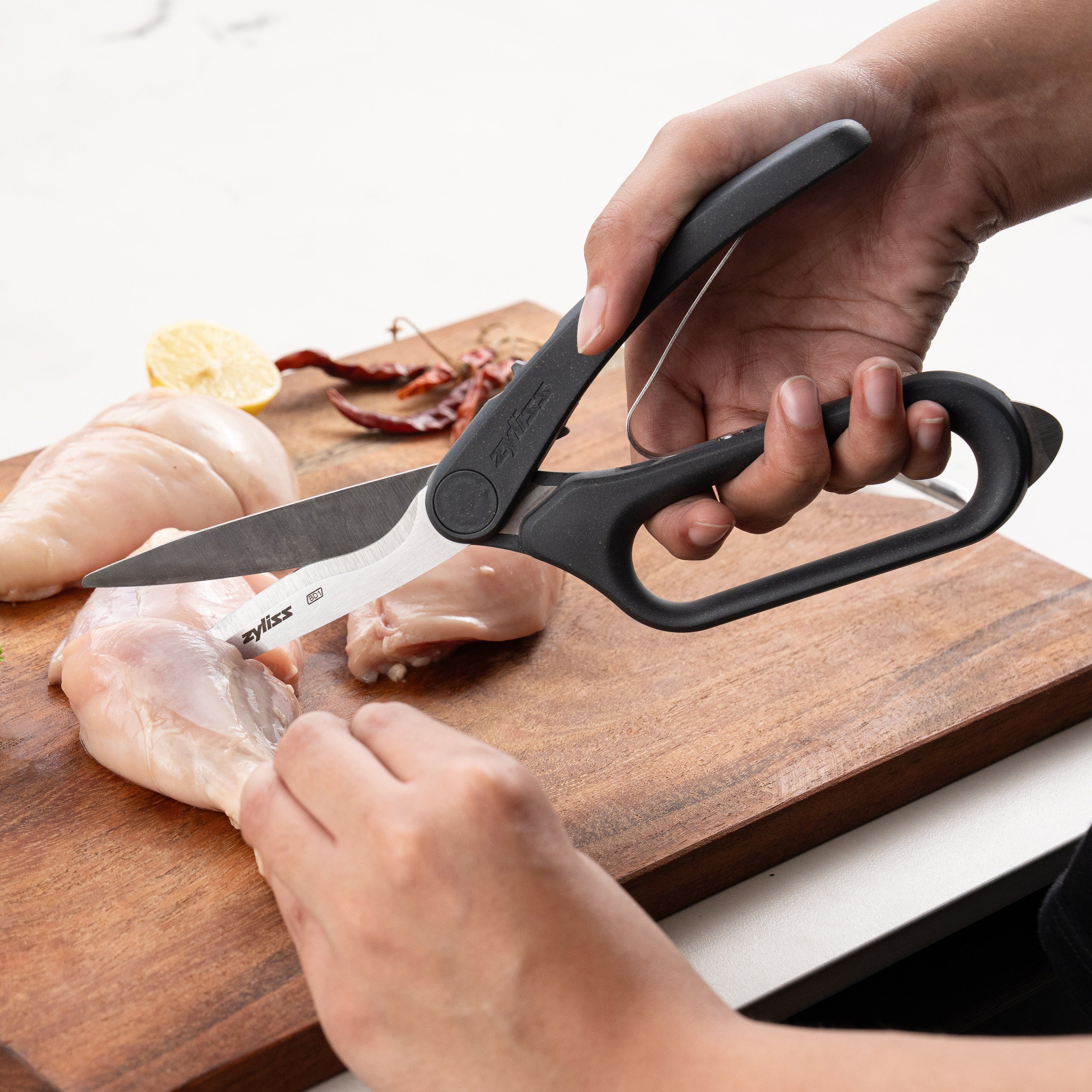 Using Zyliss All Purpose Shears with ergonomic handles and stainless steel blades, a person cuts raw chicken breast on a wooden cutting board, with lemon slice and dried chilies close by.