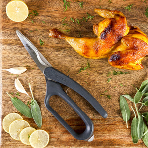 A pair of Zyliss All Purpose Shears with ergonomic handles, cooked chicken leg quarters, lemon slices, garlic cloves, sage leaves, and herbs rest on a wooden cutting board.