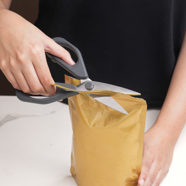 A person uses Zyliss Household Scissors Basic with stainless steel blades to cut open the top of a sealed golden-yellow plastic pouch on a white surface.