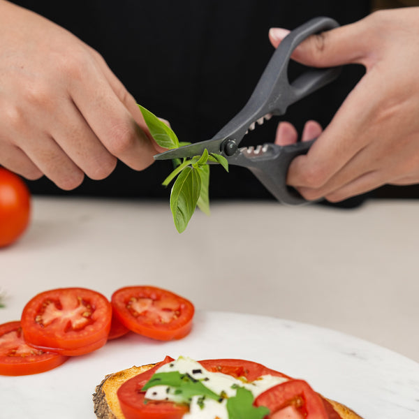 A person uses Zyliss Herb Snippers with stainless steel blades to cut fresh basil leaves over tomato and cheese-topped bread, with sliced and whole tomatoes nearby.