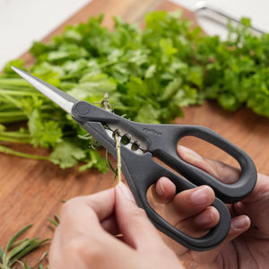 A person uses Zyliss Herb Snippers with stainless steel blades to cut fresh herbs on a wooden cutting board, with additional herbs visible in the background.