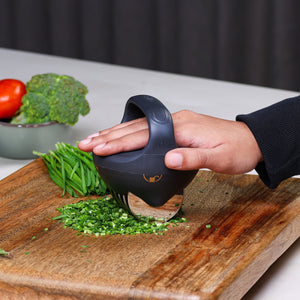 A hand uses the Zyliss Fast Cut Herb Tool, featuring stainless steel cutting wheels, to dice green herbs on a wooden cutting board. Fresh tomatoes and broccoli are visible in a bowl in the background.