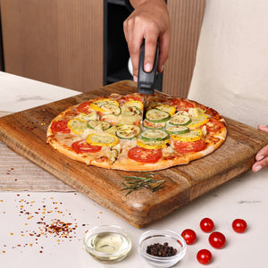 A person slices a vegetable pizza topped with tomatoes, zucchini, and herbs on a wooden board using the Zyliss Pizza Wheel. Nearby are bowls of cherry tomatoes, peppercorns, oil, and rosemary.
