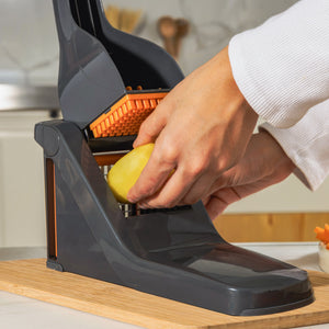 A person uses the Zyliss Potato & Vegetable Chipper, featuring stainless steel blades and a black and orange design, to slice a peeled potato on a wooden surface in the kitchen.