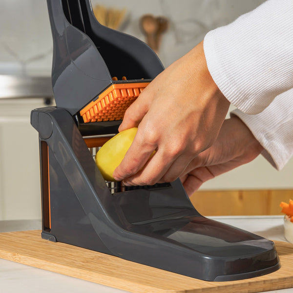 A person uses the Zyliss Potato & Vegetable Chipper, featuring stainless steel blades and a black and orange design, to slice a peeled potato on a wooden surface in the kitchen.