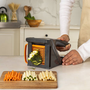 Using the Zyliss Potato & Vegetable Chipper, a person cuts zucchini into sticks. On the kitchen table, neatly arranged carrot, zucchini, and parsnip sticks rest on a wooden board.