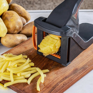 A potato is being sliced into thin strips with the Zyliss Potato & Vegetable Chipper, featuring stainless steel blades and a black-orange design, on a wooden cutting board with whole and peeled potatoes nearby.