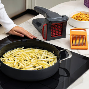 A person stands by a stove frying sliced potatoes as the Zyliss Potato & Vegetable Chipper, with stainless steel blades, a grater, and a bowl of cooked fries rest on the counter; the chipper is dishwasher safe.