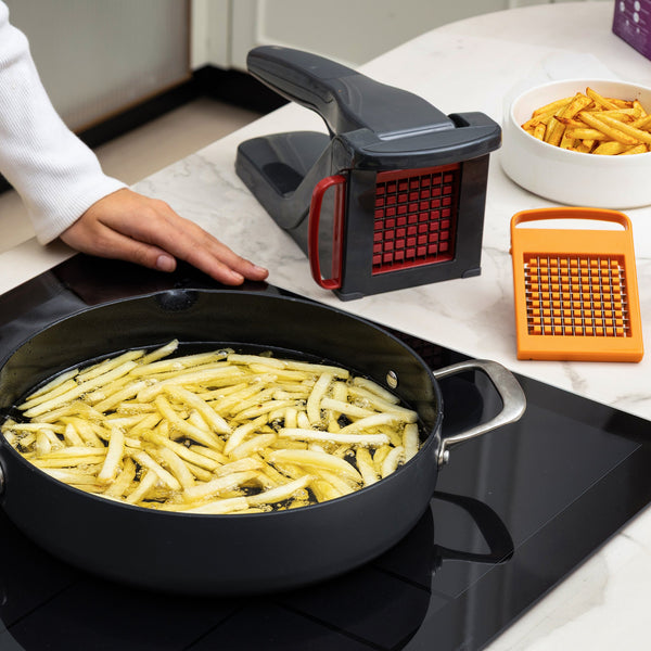 A person stands by a stove frying sliced potatoes as the Zyliss Potato & Vegetable Chipper, with stainless steel blades, a grater, and a bowl of cooked fries rest on the counter; the chipper is dishwasher safe.