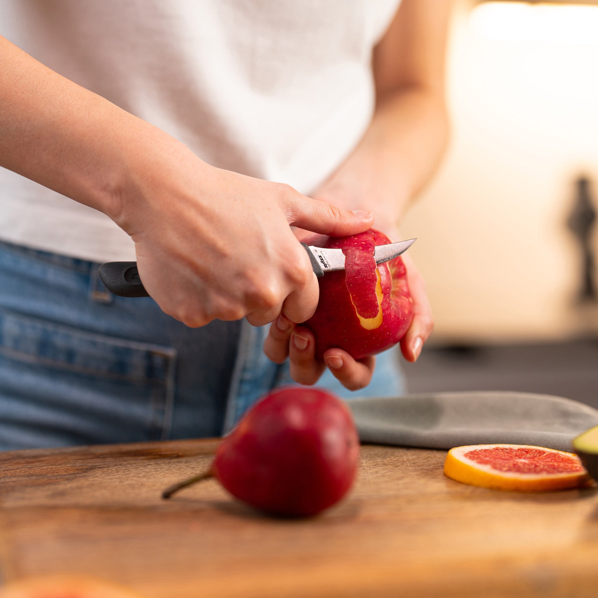A person in a white shirt and jeans is peeling a red pear with a small knife over a wooden cutting board. Another red pear and a slice of citrus fruit are also on the board.