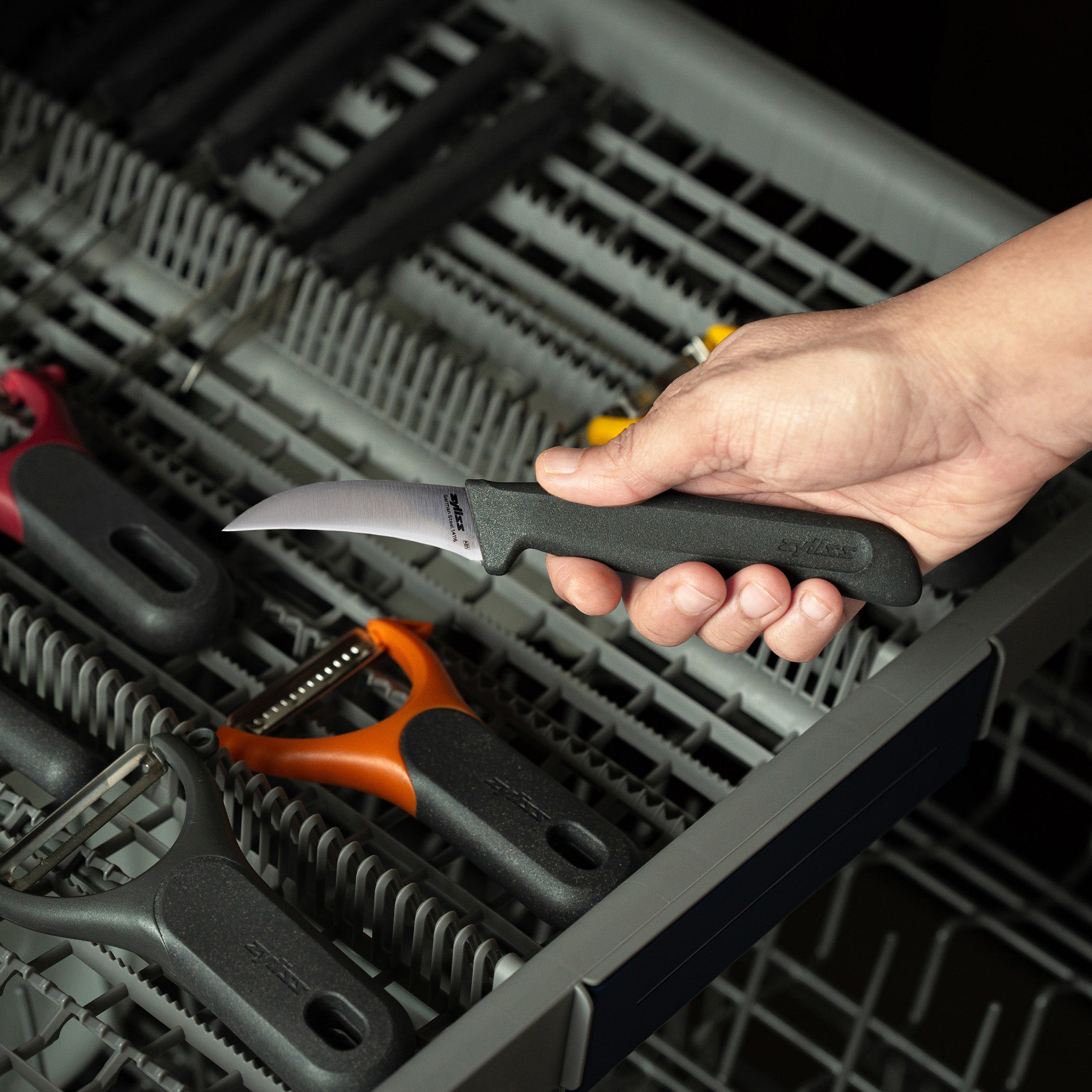 A hand is placing a small paring knife with a black handle into a dishwasher rack, alongside other kitchen utensils like peelers.