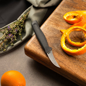 A Zyliss Peeling Knife 6cm / 2½ with an ergonomic handle sits on a wooden cutting board beside orange peels, while an unpeeled orange and dried herbs are nearby on a gray surface. 