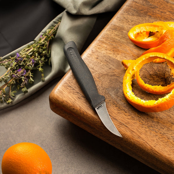 A Zyliss Peeling Knife 6cm / 2½ with an ergonomic handle sits on a wooden cutting board beside orange peels, while an unpeeled orange and dried herbs are nearby on a gray surface. 