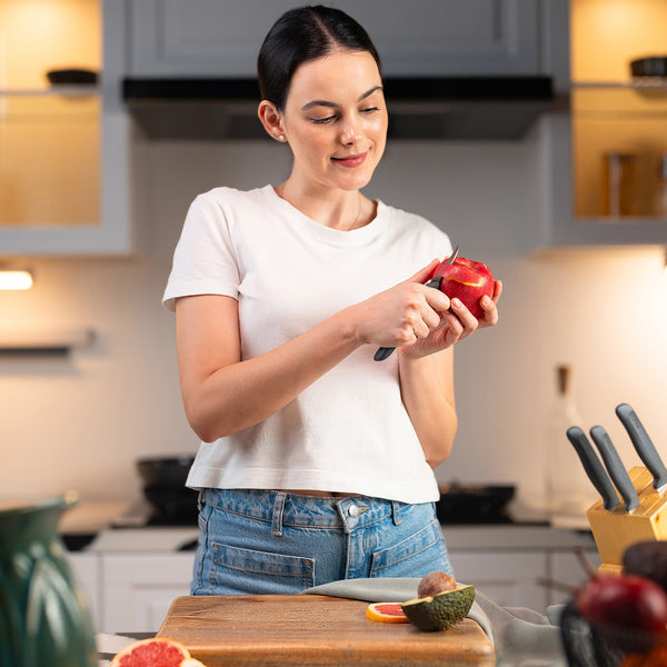 A woman in a white tee and jeans peels a red apple with the Zyliss Peeling Knife 6cm/2½, which has an ergonomic handle, in a modern kitchen beside a wooden cutting board filled with assorted fruits. 