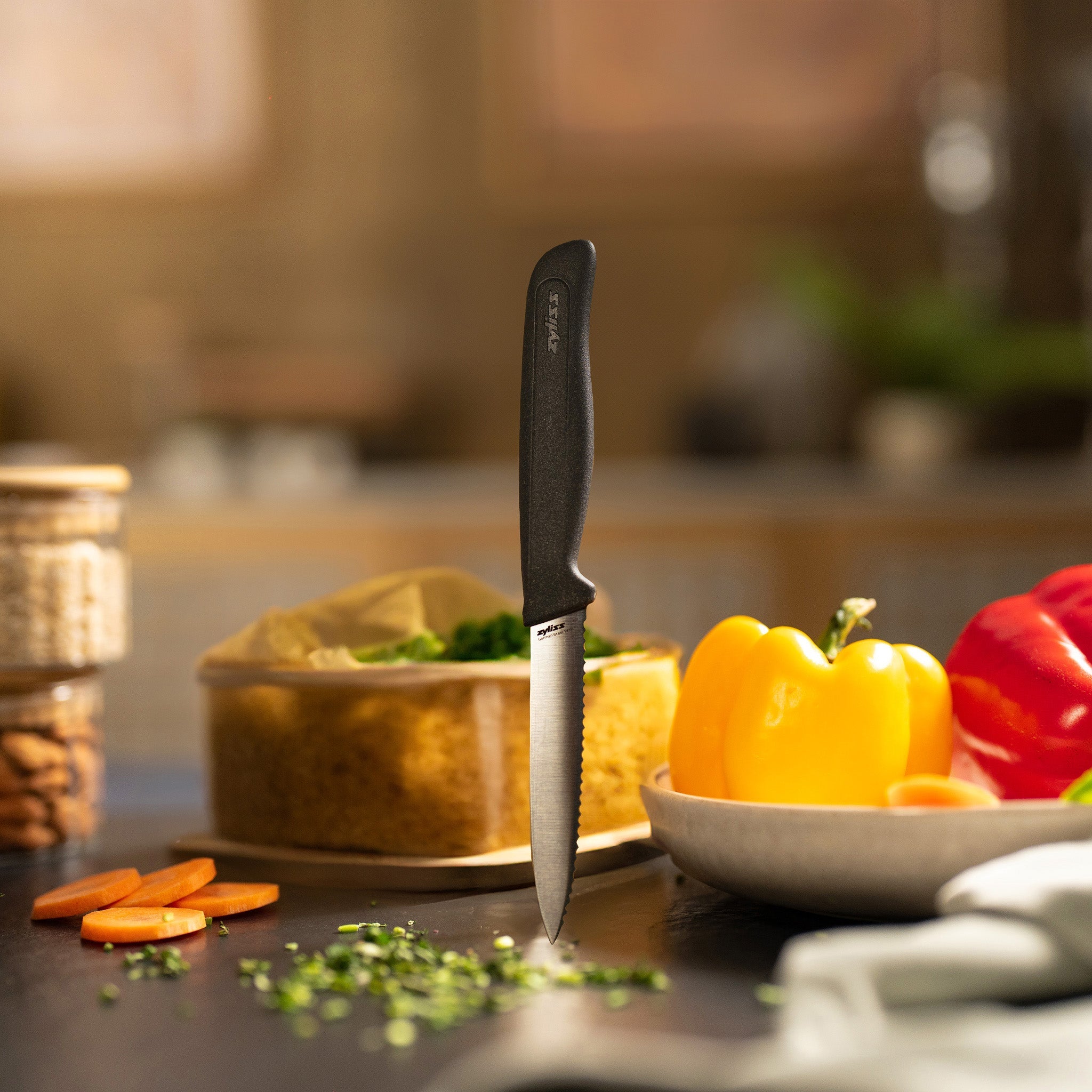 A Zyliss Paring Knife Serrated 10cm / 4 with a black handle stands upright on a kitchen counter, surrounded by chopped herbs, sliced carrots, yellow and red bell peppers, a plate, and storage containers. The background is softly blurred. 